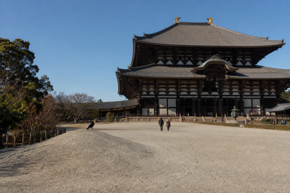 A wide gravel clearing in front of a large wooden temple hall under a blue sky