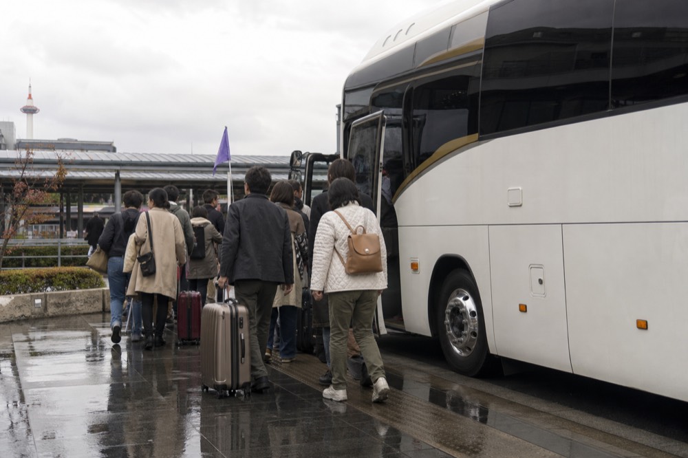 A modern coach parked outside Kyoto Station with passengers boarding and a guide holding a flag