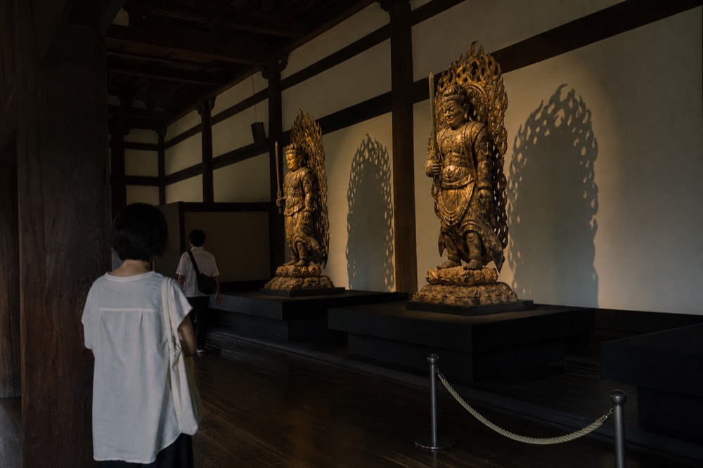A quiet gallery hall with wooden Buddhist statues displayed on plinths under soft light